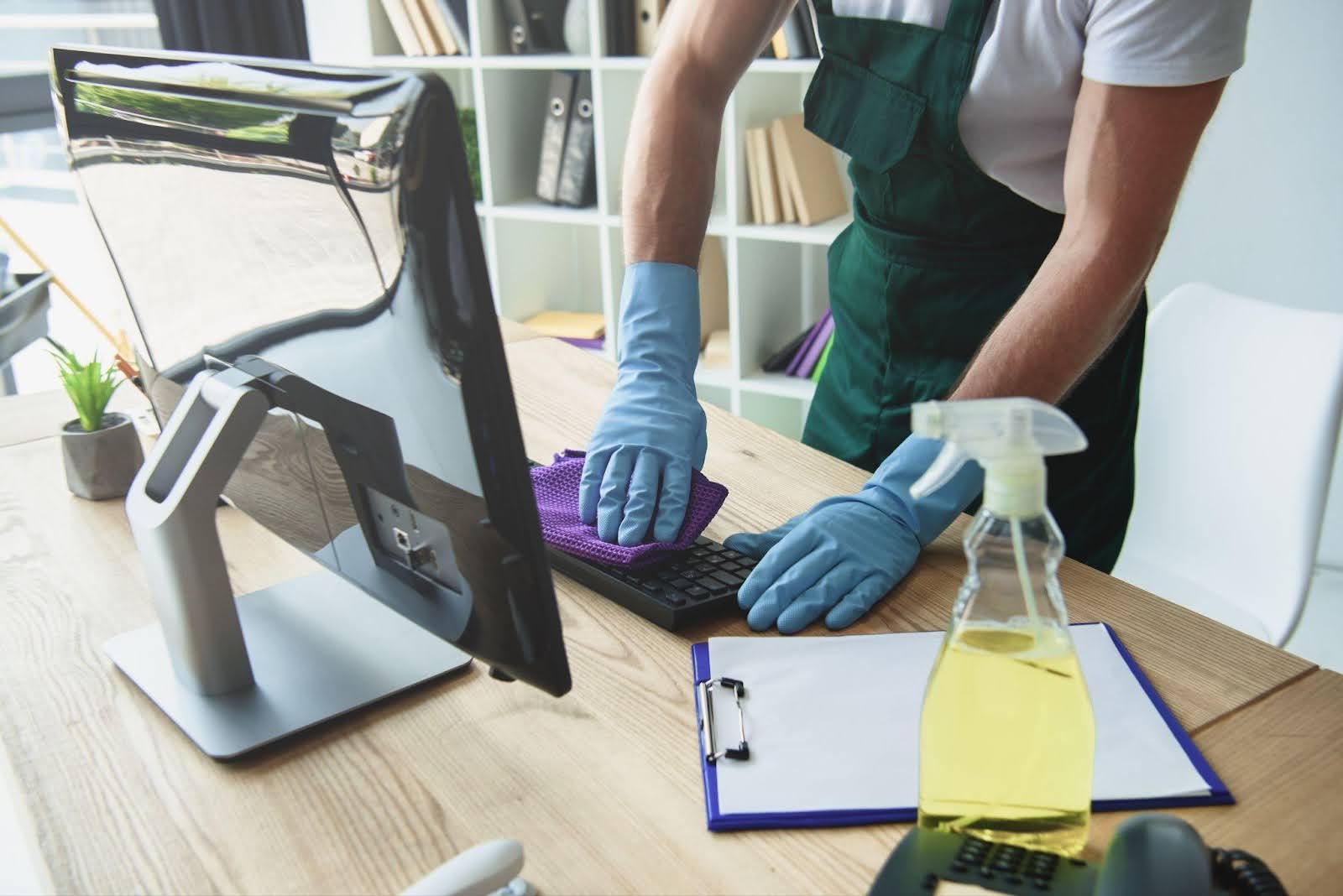 A person in gloves cleans a computer keyboard with a purple cloth in an office