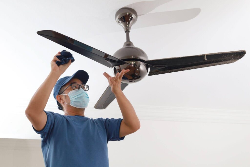 A person wearing a mask and glasses cleans a ceiling fan with a cloth