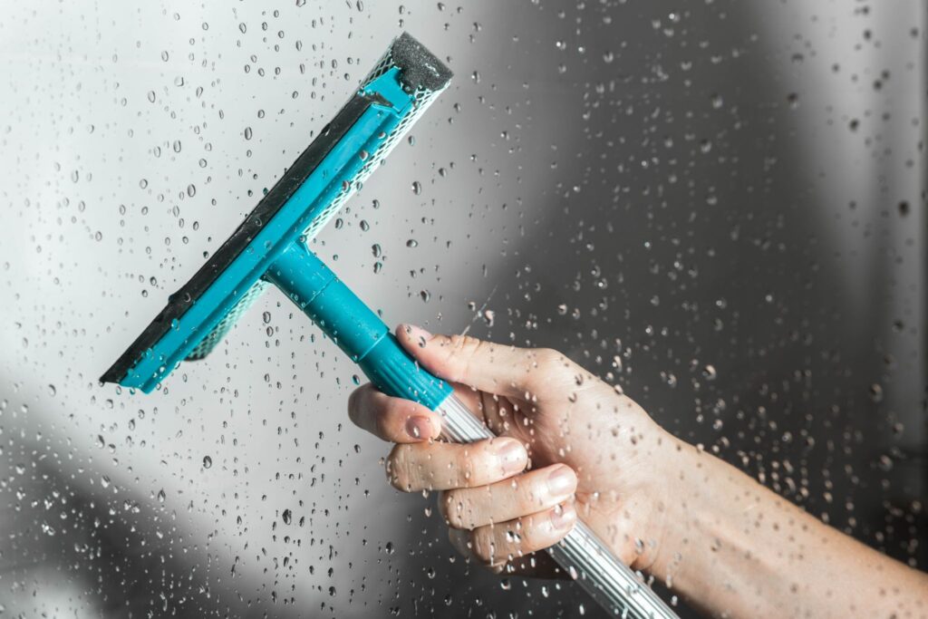 A hand holding a teal and black squeegee against a glass surface covered in water droplets.