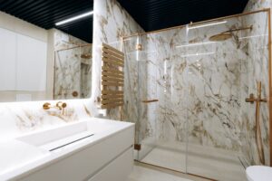 A modern, luxury bathroom featuring white marble walls with gold veining, a sleek white vanity with a gold faucet, and a spacious glass-enclosed walk-in shower with gold fixtures.