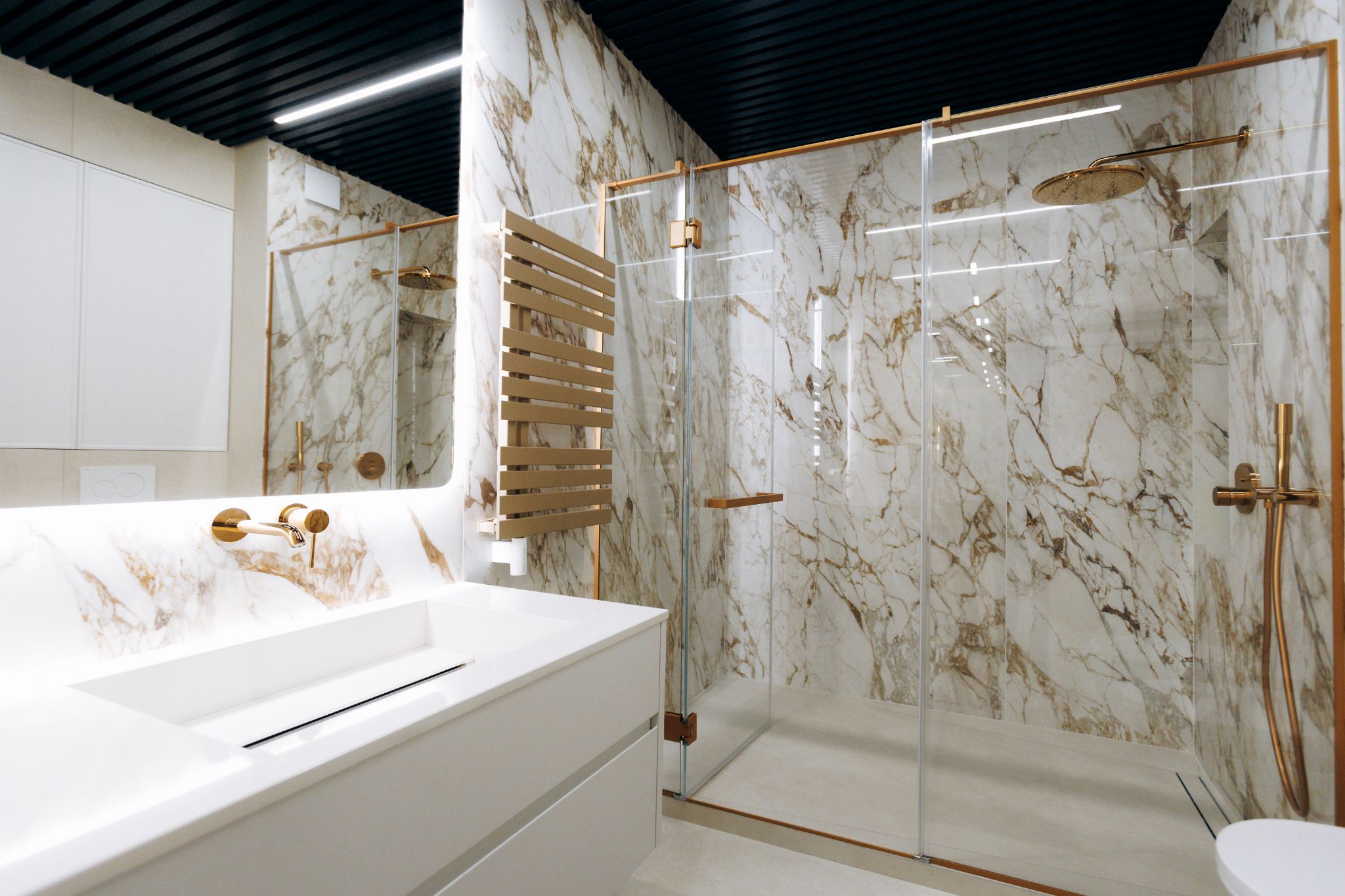 A modern, luxury bathroom featuring white marble walls with gold veining, a sleek white vanity with a gold faucet, and a spacious glass-enclosed walk-in shower with gold fixtures.