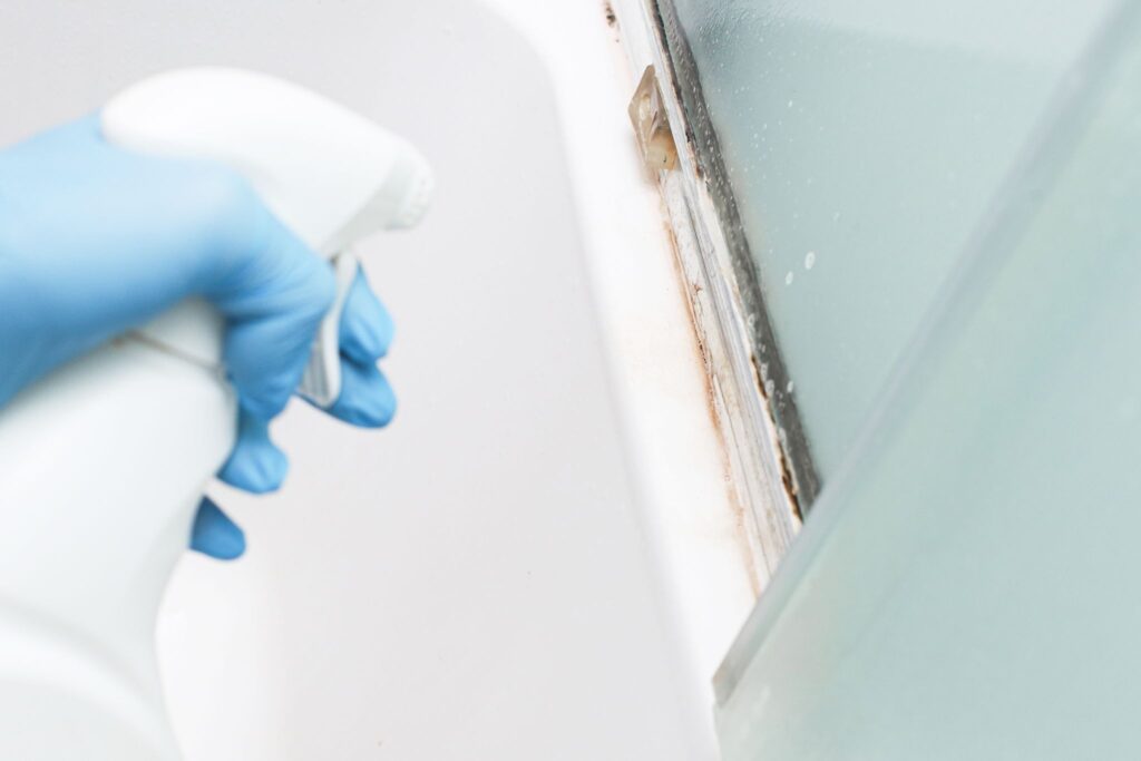 A person wearing a blue glove sprays a white cleaning bottle onto a dirty shower track covered in orange-brown grime and mildew.