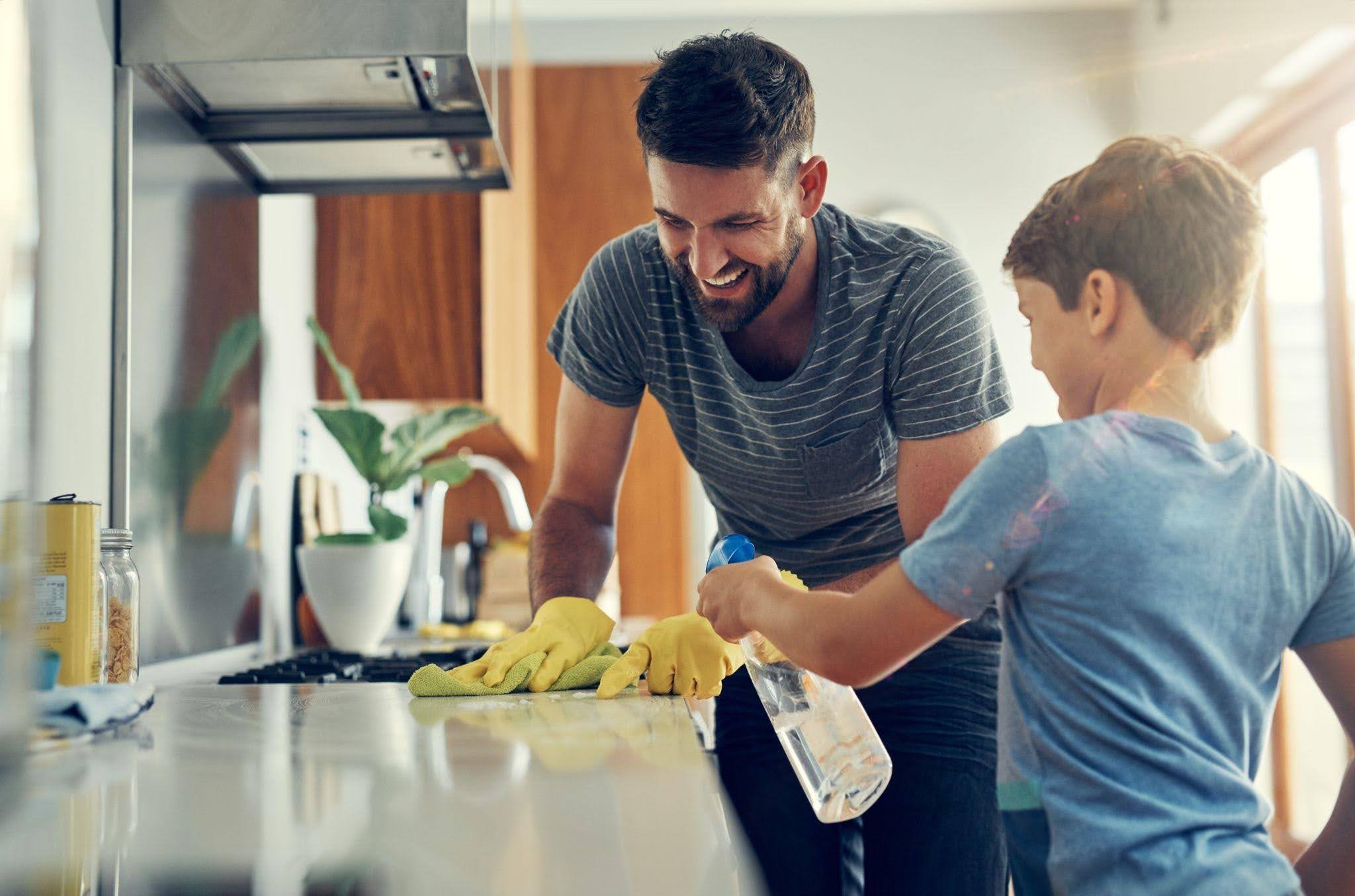 A man and a boy are collaboratively cleaning the kitchen counter using cloths and cleaning supplies