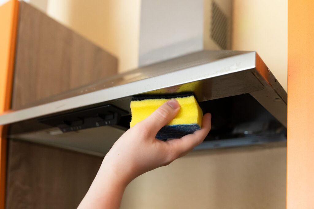 Hand using a yellow scrubbing sponge to clean the underside of a stainless steel kitchen range hood.