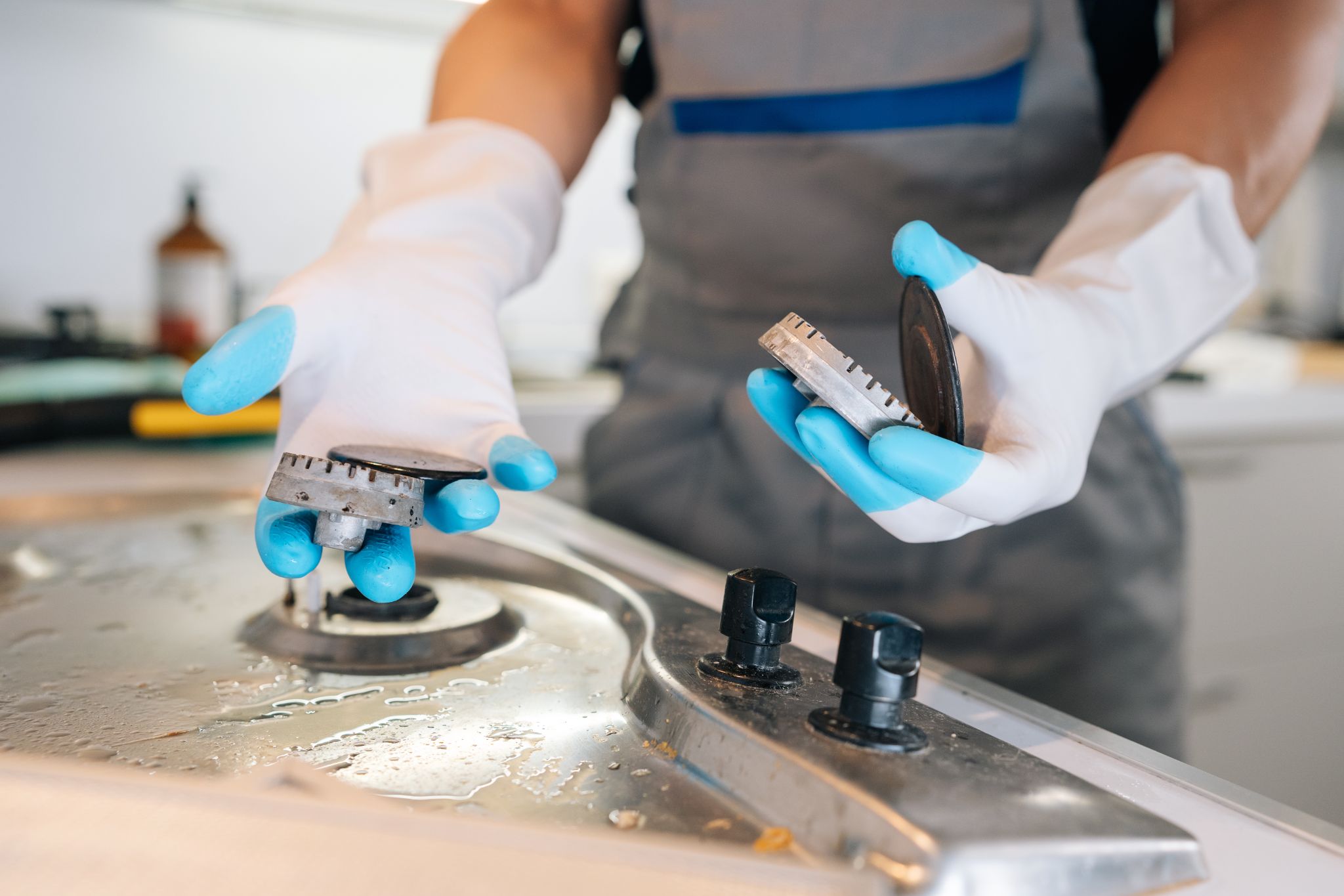 Person in protective gloves holding detached burner caps while cleaning a stainless steel gas stovetop.