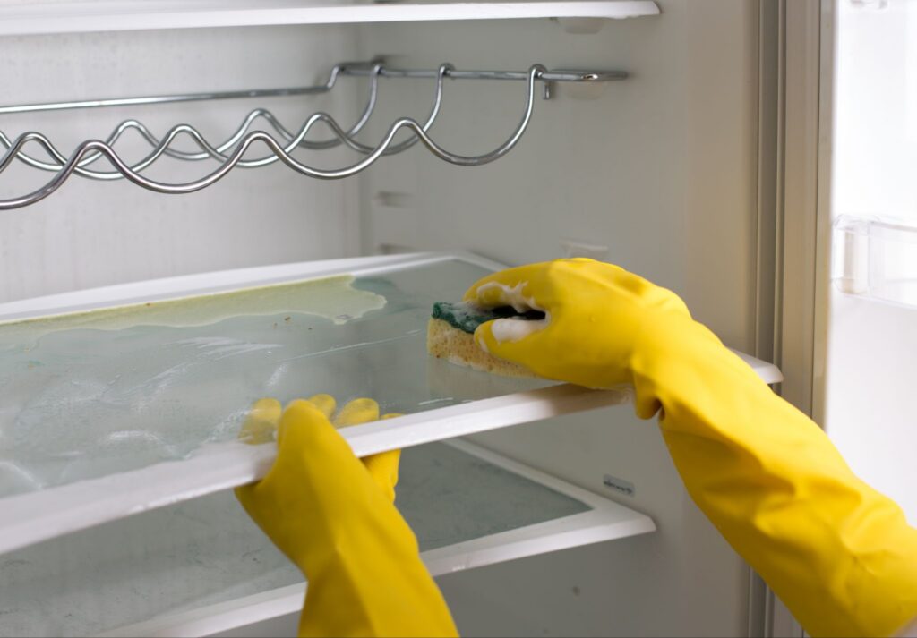 Person in yellow rubber gloves scrubbing a dirty glass shelf inside an empty refrigerator with a soapy sponge.